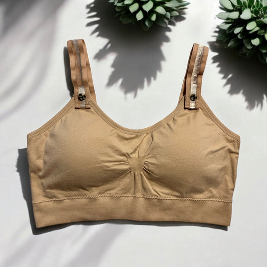 The photo displays a flat lay of a nude bra which closely resembles a sports bra. The straps are nude on the sides and clear within the middle. To promote easy access the straps are equipped with buttons on the front to slide the straps into. Three succulents are styled around the nude bra.