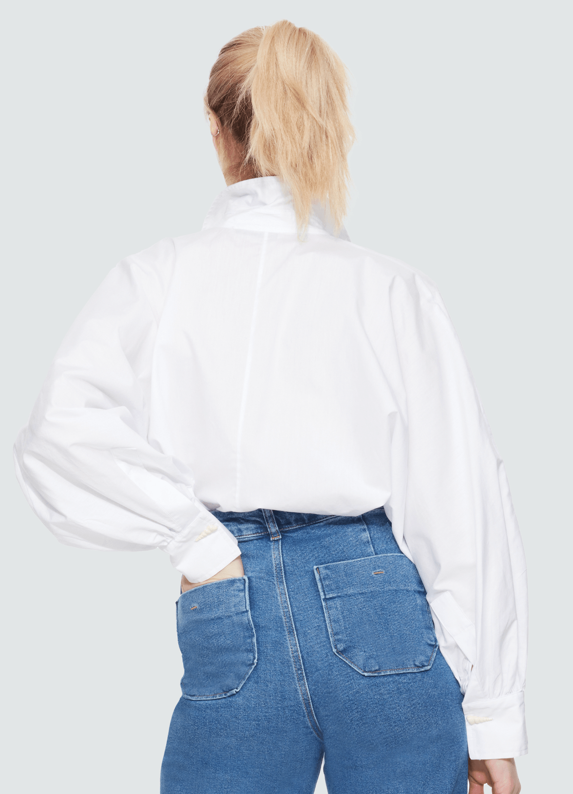 Woman in her 30s with blonde hair is, facing the back wall and  wearing a crisp white button up blouse that entails a larger flattering sleeve - which are pleated at the wrist. To complement the blouse, the woman is wearing high waist jeans with her left hand in her back left pocket.