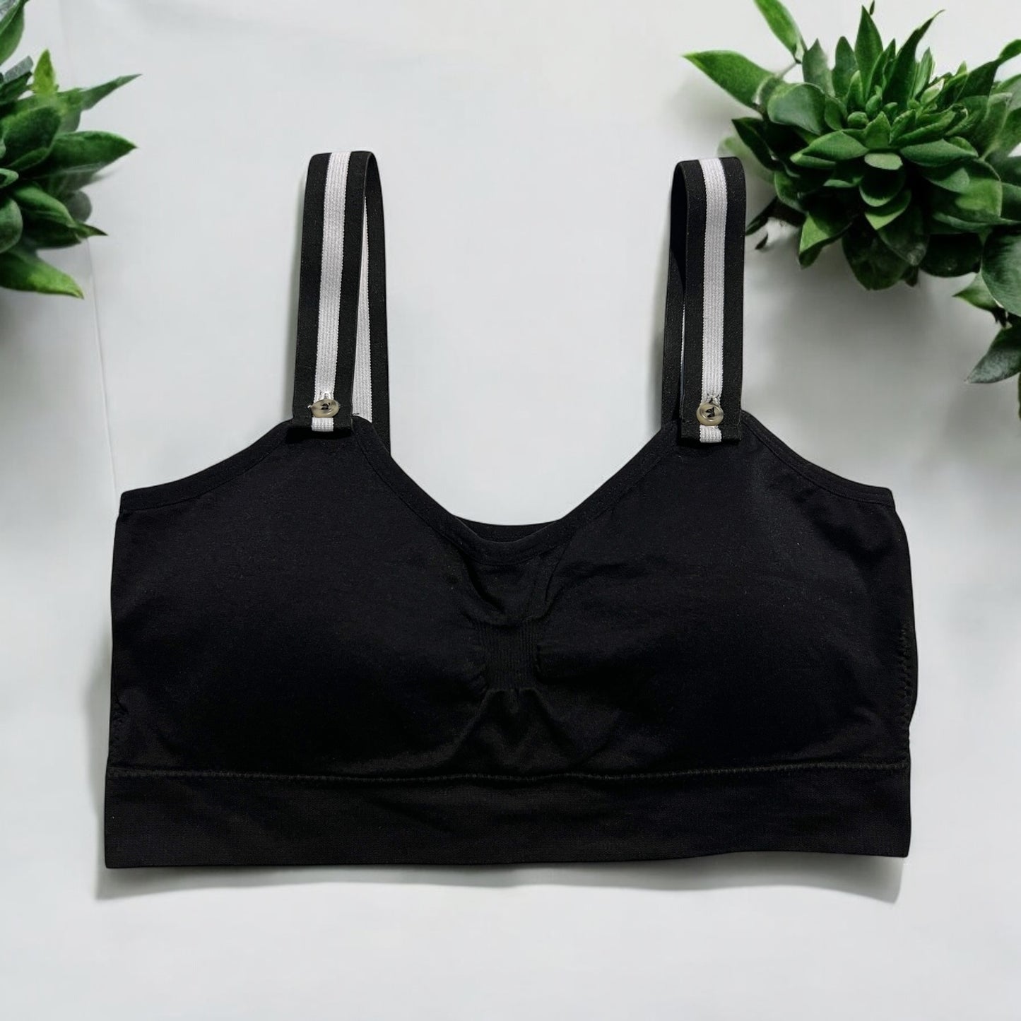 The photo displays a flat lay of a black bra which closely resembles a sports bra. The straps are black and white. To promote easy access the straps are equipped with buttons on the front to slide the straps into. Succulents are styles around the black bra.