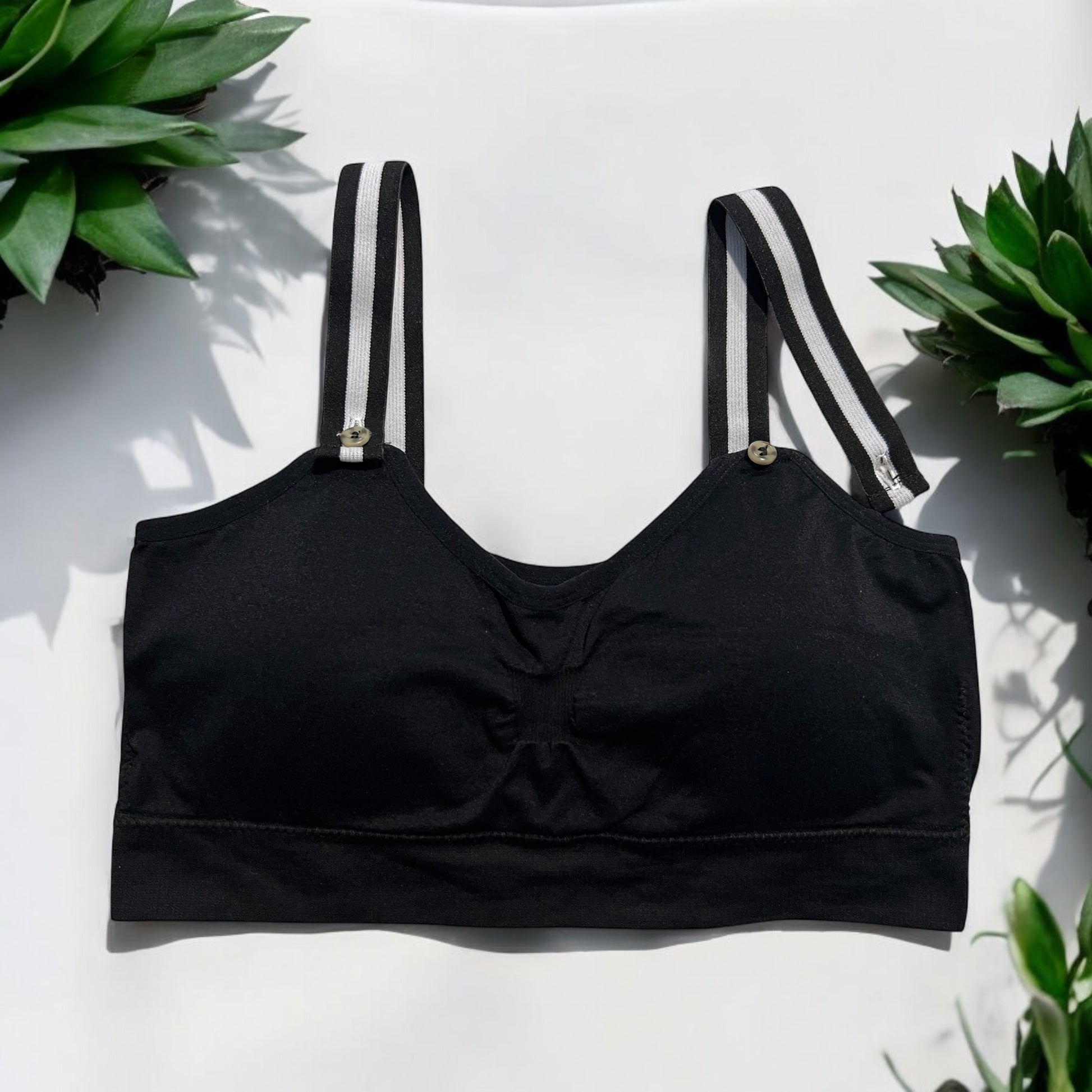 The photo displays a flat lay of a black bra which closely resembles a sports bra. The straps are black and white. To promote easy access the straps are equipped with buttons on the front to slide the straps into. Succulents are styles around the black bra.