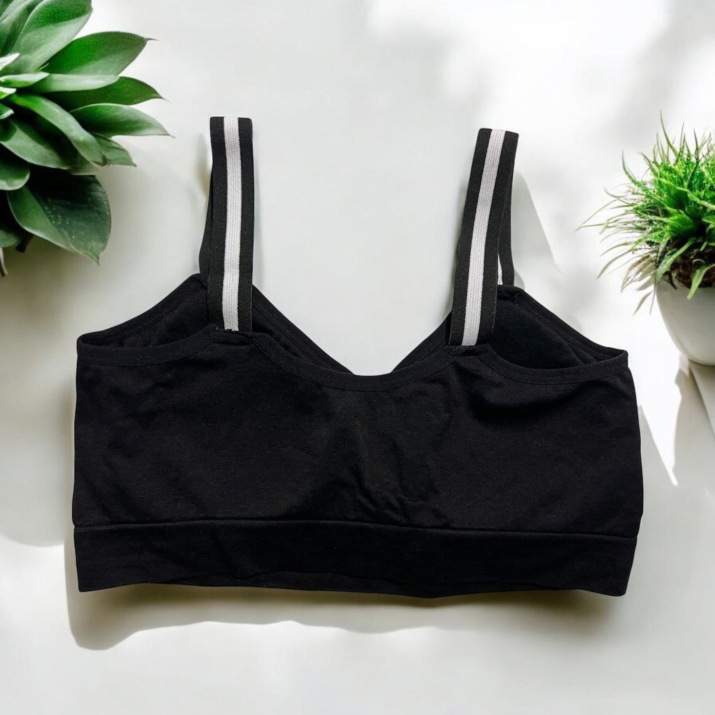 The photo displays a flat lay of the back of the black bra which closely resembles a sports bra. The straps are black and white. To promote easy access the straps are equipped with buttons on the front to slide the straps into. Succulents are styles around the black bra.