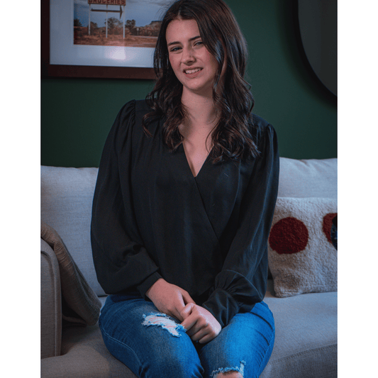 Young female with brown hair is wearing a loose drape blouse with flowing sleeves that button at the wrist. To complement the black top, the female is wearing jeans with slight rips. The female is sitting on a soft gray couch.