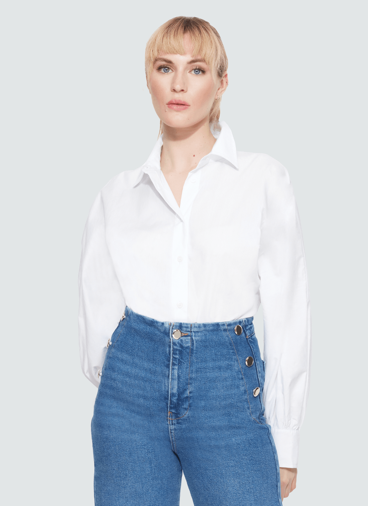 Woman in her 30s with blonde hair is wearing a crisp white button up blouse that entails a larger flattering sleeve. To complement the blouse, the woman is wearing high waist jeans.