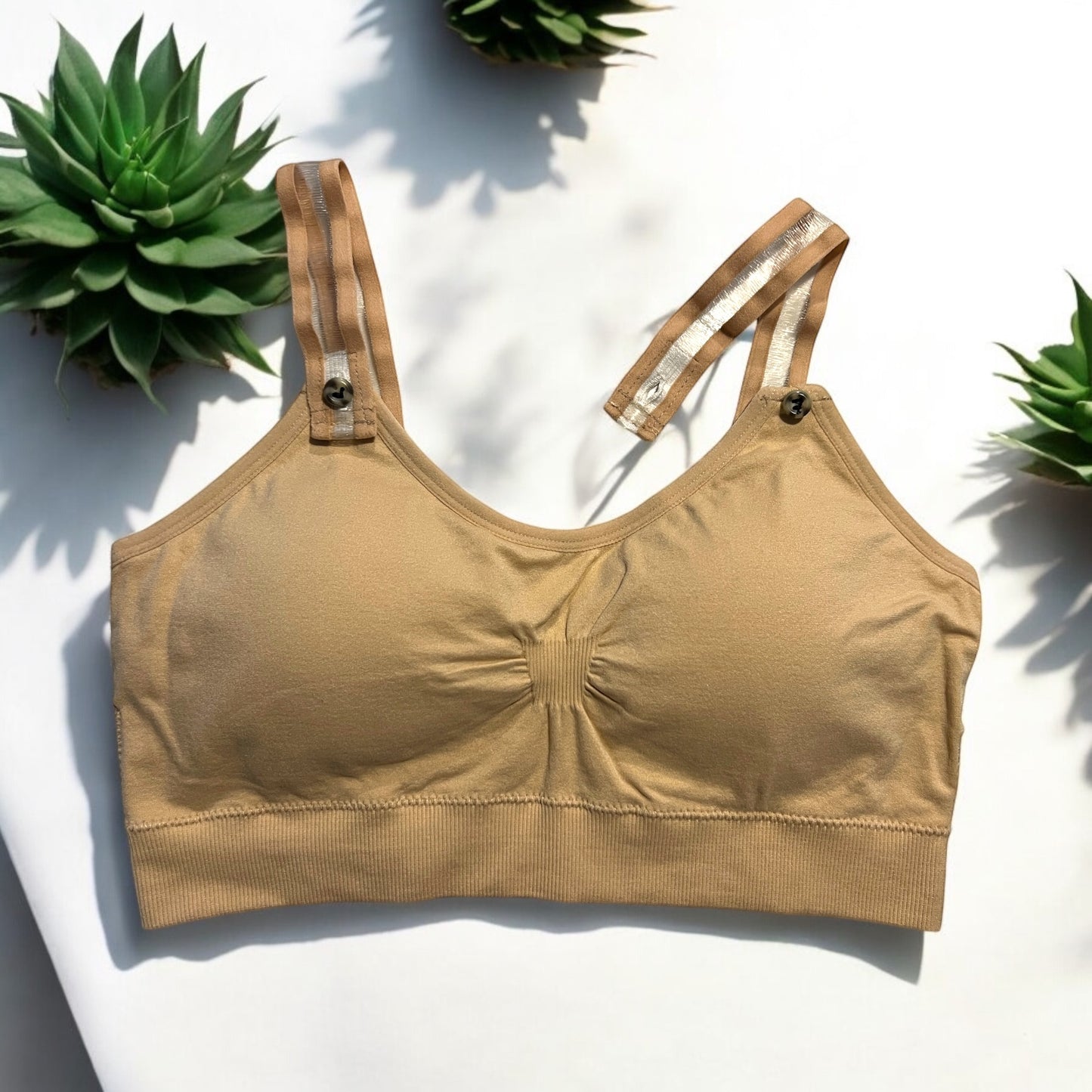 The photo displays a flat lay of a nude bra which closely resembles a sports bra. The straps are nude on the sides and clear within the middle. To promote easy access the straps are equipped with buttons on the front to slide the straps into. Three succulents are styles around the nude bra.